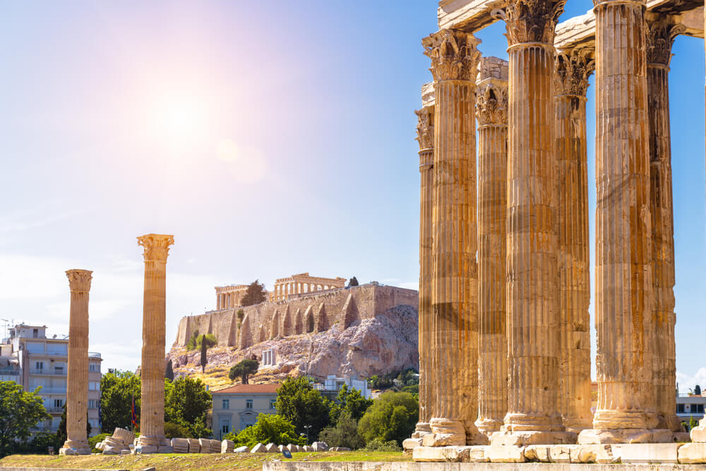 Vista soleggiata del Tempio di Zeus con l'Acropoli di Atene, Grecia. Iconici punti di riferimento con antiche rovine e imponenti colonne. Esplora la storia in un viaggio unico.